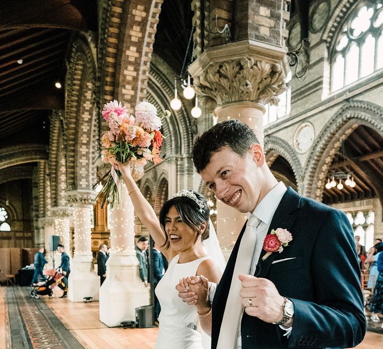 Groom wears colourful floral buttonhole alongside his bride who lifts her matching bouquet in the air 