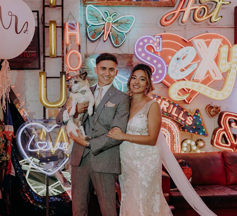 Bride in sleeveless plunge lace wedding dress  and cathedral length embroidered veil standing with groom in grey three piece wedding suit with yellow suit and yellow pocket square and their little puppy 