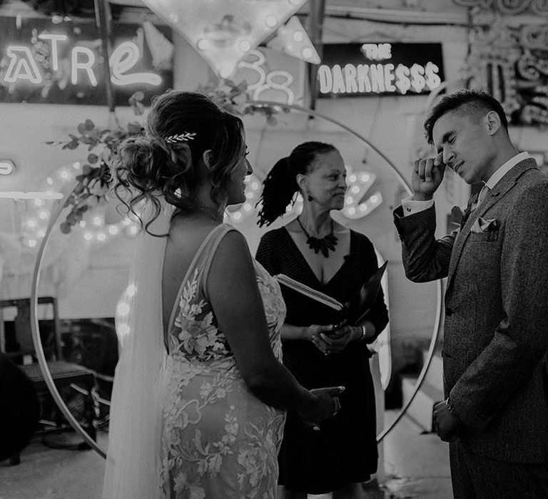 Groom in three piece grey suit wiping tear from his eye with bride in lace open back detail wedding dress and celebrant 