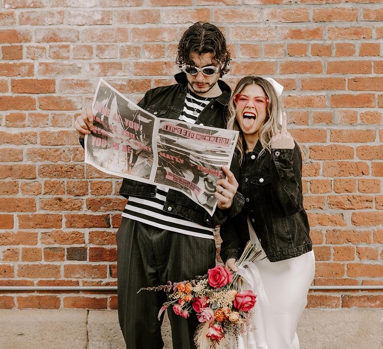 Bride in v neck satin wedding dress and black denim jacket holding pink rose, pampas grass and foliage bouquet with groom in black denim jacket, black and white striped long sleeve t shirt and grey pinstripe trousers reading tabloid-style newspaper wedding stationery 