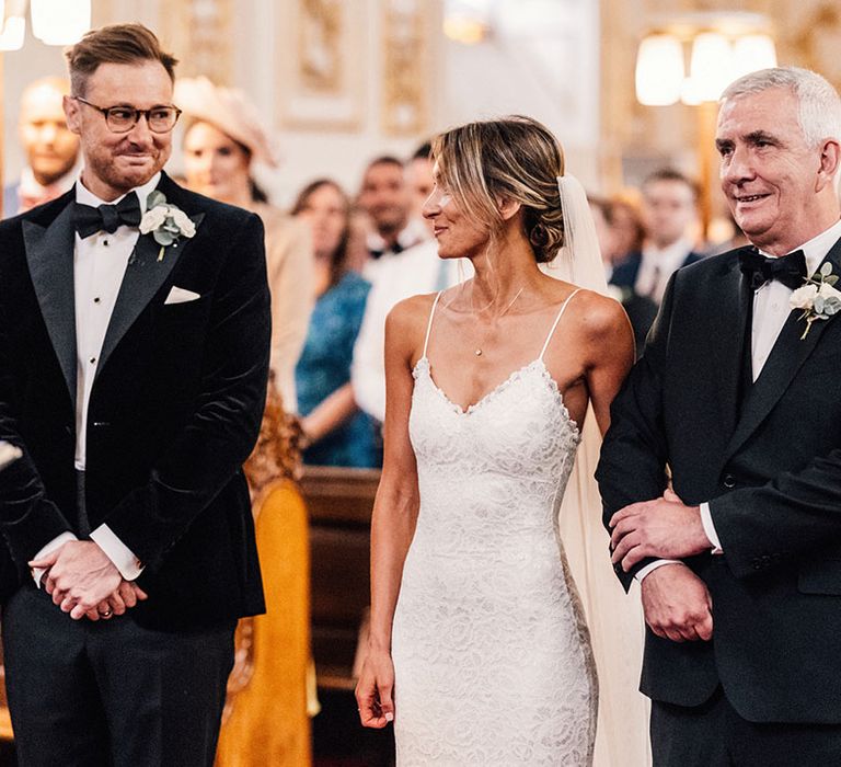 Father of the bride in a black tuxedo stands at the altar with the bride in a lace wedding dress and the groom in matching black tux for the church wedding ceremony