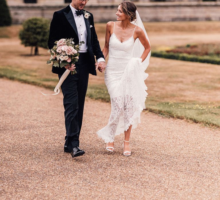 The groom in glasses wears a traditional black velvet tuxedo walking with the bride in a  subtle mermaid style lace fit and flare wedding dress 