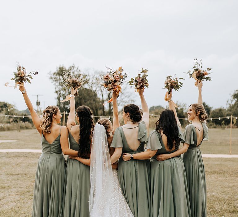 The bridesmaids hold small autumnal bouquets in light green bridesmaid dresses with the bride for fun bridal party photos 