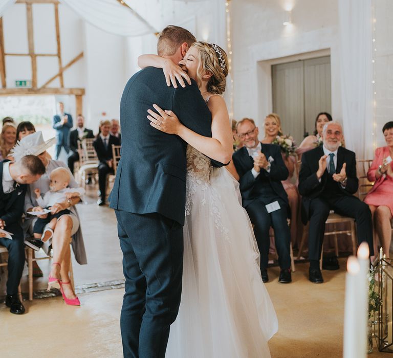 Bride with blonde braided hairstyle with sparkly headpiece embraces the groom in a navy suit at the altar in front of the wedding guests 