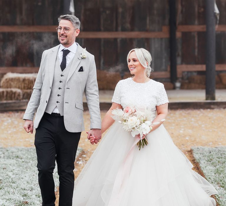 Bride in big princess tulle skirt with short sleeve top walking hand in hand with the groom in light grey suit jacket and waistcoat 