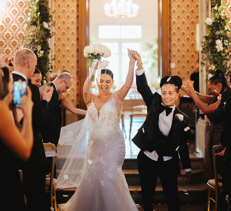 Bride in black snakeskin print tuxedo with black bowtie and white rose boutonniere and bride in lace sleeveless v neck Enzoani wedding dress with tulle finish and church length veil holding white garden rose bridal bouquet doing confetti exit at black-tie lesbian wedding at Grittleton House