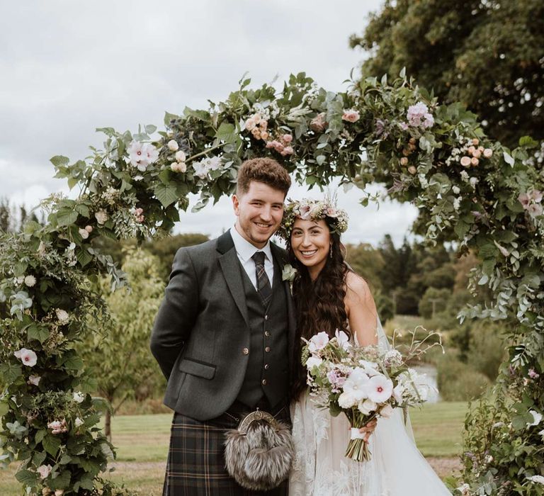 Groom in dark grey blazer and waistcoat, dark tartan tie with white flower boutonniere, dark grey and blue tartan kilt and wedding sporran standing with bride in celestial 3d appliqué sheer overlay wedding dress and bridal flower crown holding large white rose, peony, dried flower and foliage bridal bouquet and miniature schnauzer puppy 