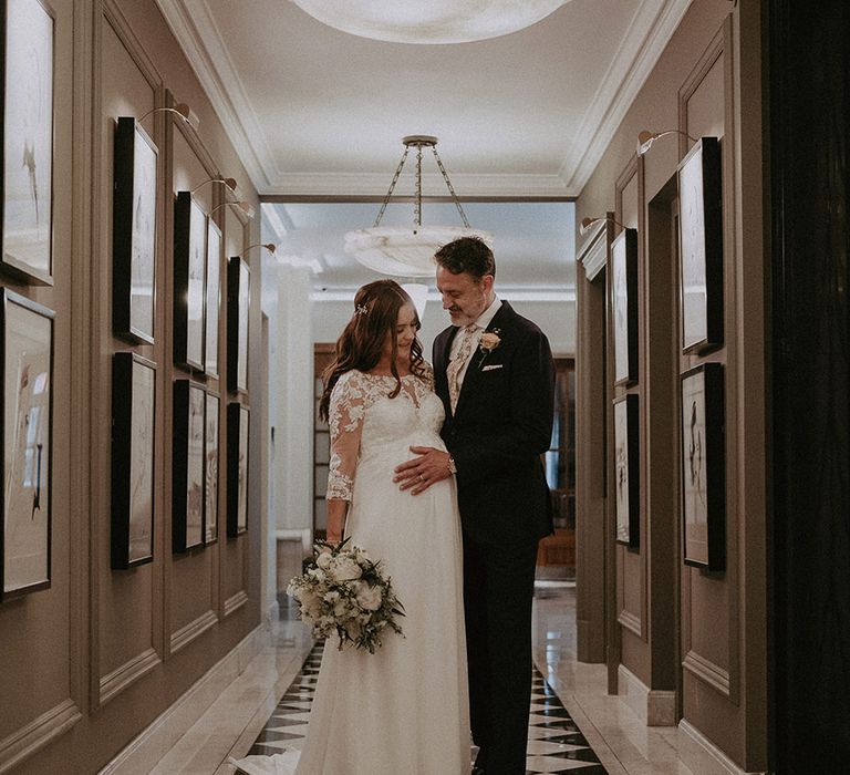 Bride in maternity wedding dress with baby bump with the groom in navy suit resting his hands on his baby bump 