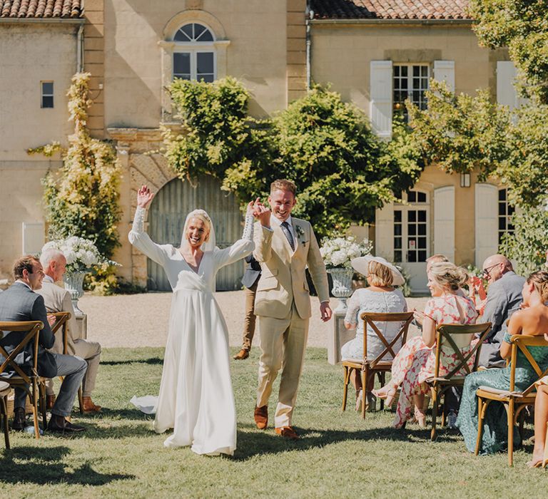 Bride in an Emma Beaumont wedding dress and groom in a beige suit descending down the aisle at Château de Malliac