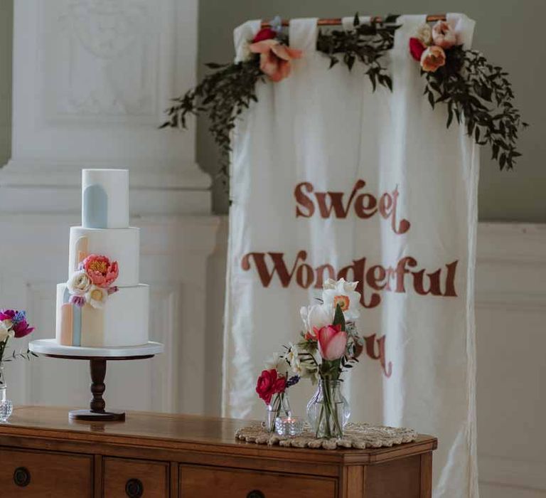 Three tiered wedding cake with pastel icing and rose decorations on wooden chest of drawers with white and red wedding banner 