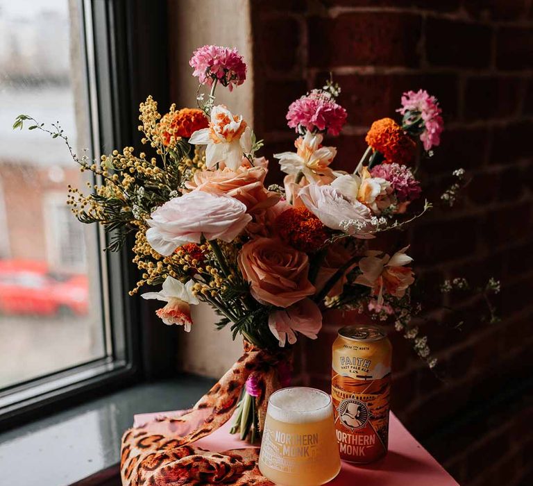 Colourful mixed flower bridal bouquet tied with leopard print ribbon on pink wedding stand with craft beer at Northern Monk Refectory Leeds