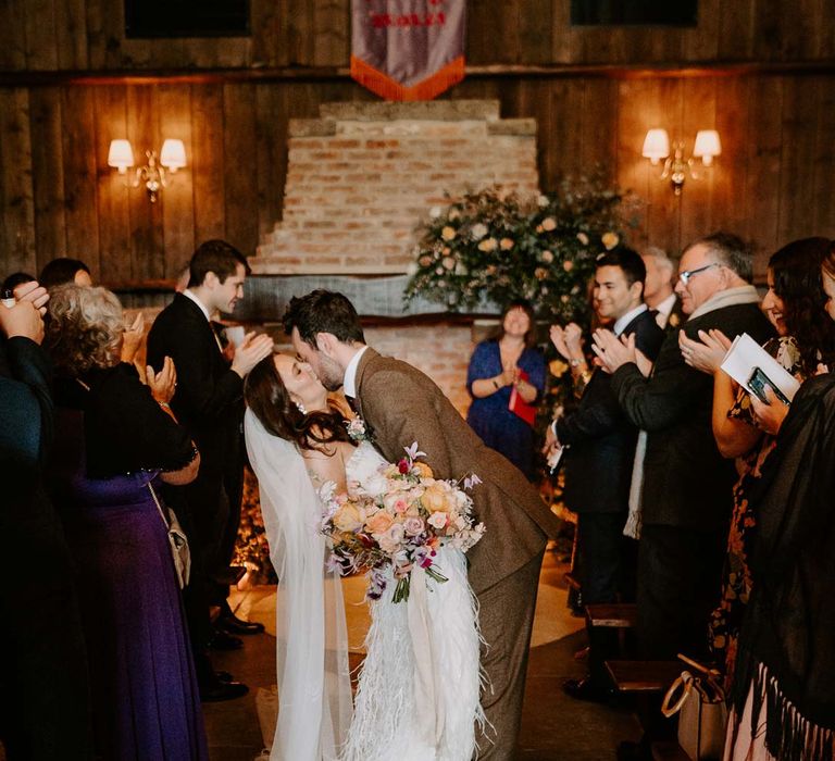 Groom in chocolate brown grooms suit kissing bride in strapless ivory fringed crepe gown and chapel length veil whilst walking down the aisle at Willow Marsh Farm Loughborough 