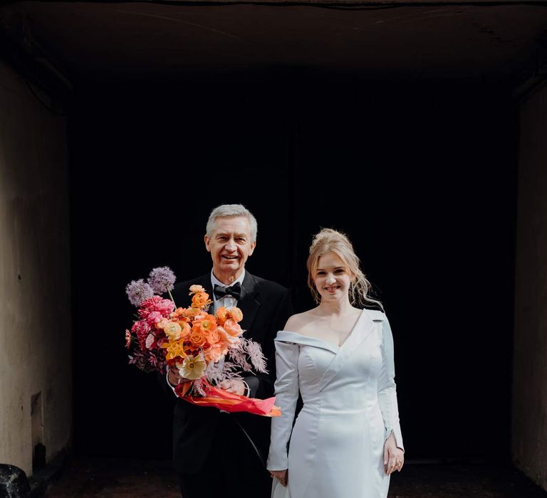 Bride in asymmetrical off shoulder long sleeve satin wedding dress standing with father of the bride holding colourful bridal bouquet with pink garden roses, purple giant alliums, dark red poppies and foliage 