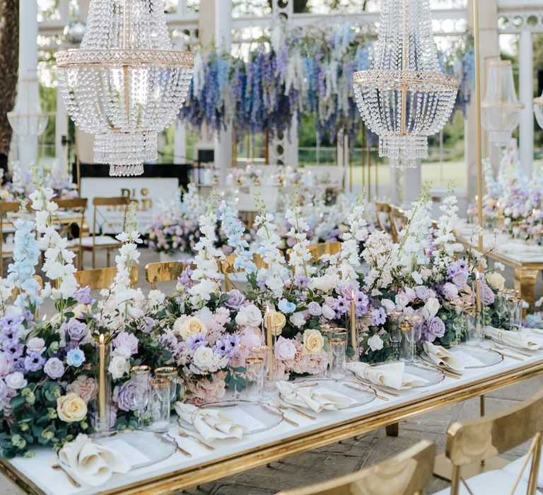 Syon Park wedding venue reception room with gold chairs, botanical tablescapes with large pastel flower centrepieces, gold tapered candles, gold rimmed tableware and suspended pastel foliage decor 