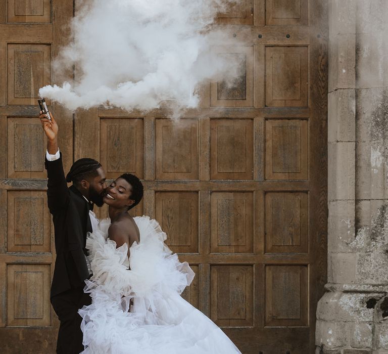 Groom in black tuxedo waving white smoke bomb with the bride in statement tulle gown 