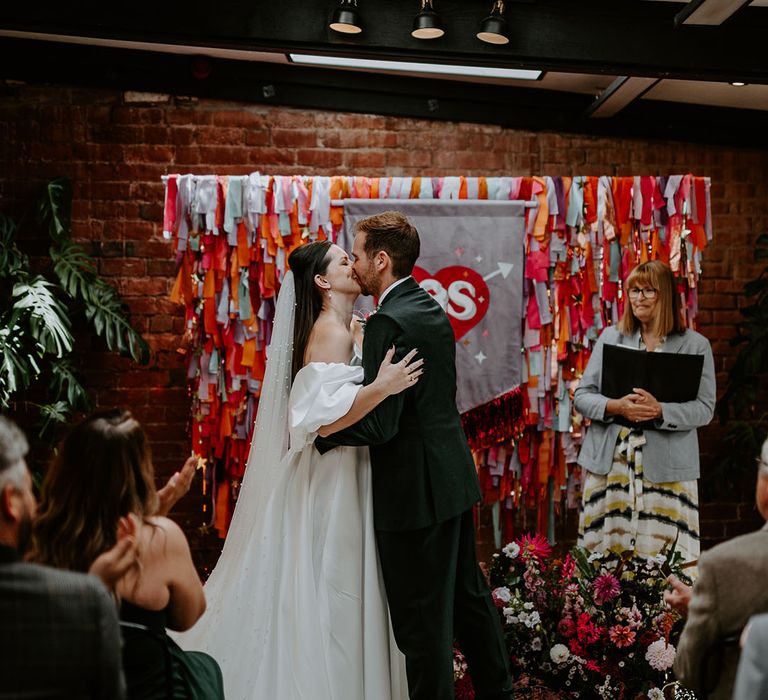 Bride wearing pearl wedding veil kissing groom for their first kiss with purple custom wedding banner 