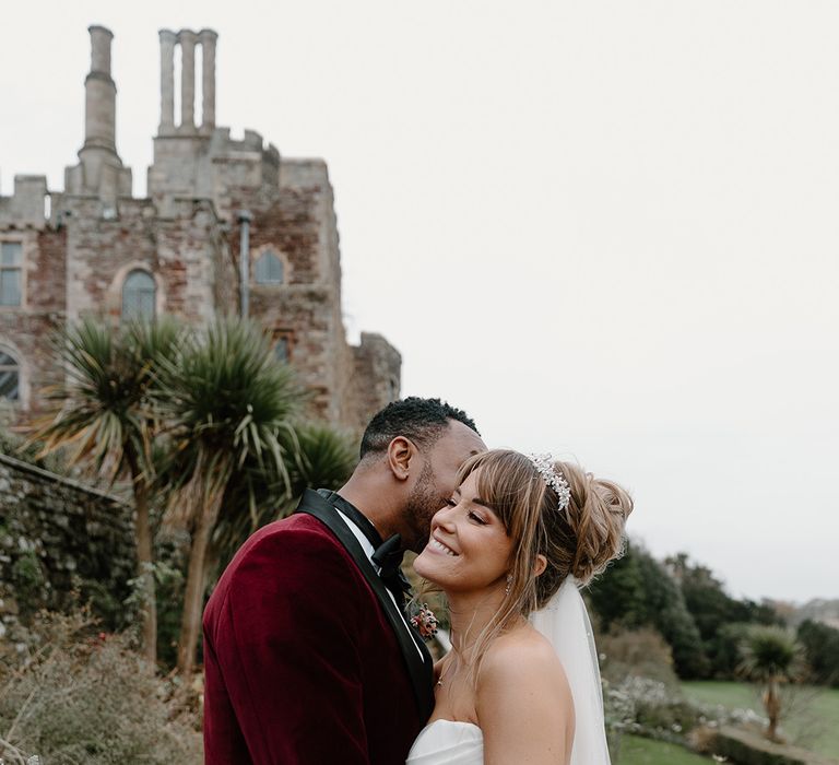 The groom in a deep red velvet suit jacket kisses the bride in a romantic couple photo 