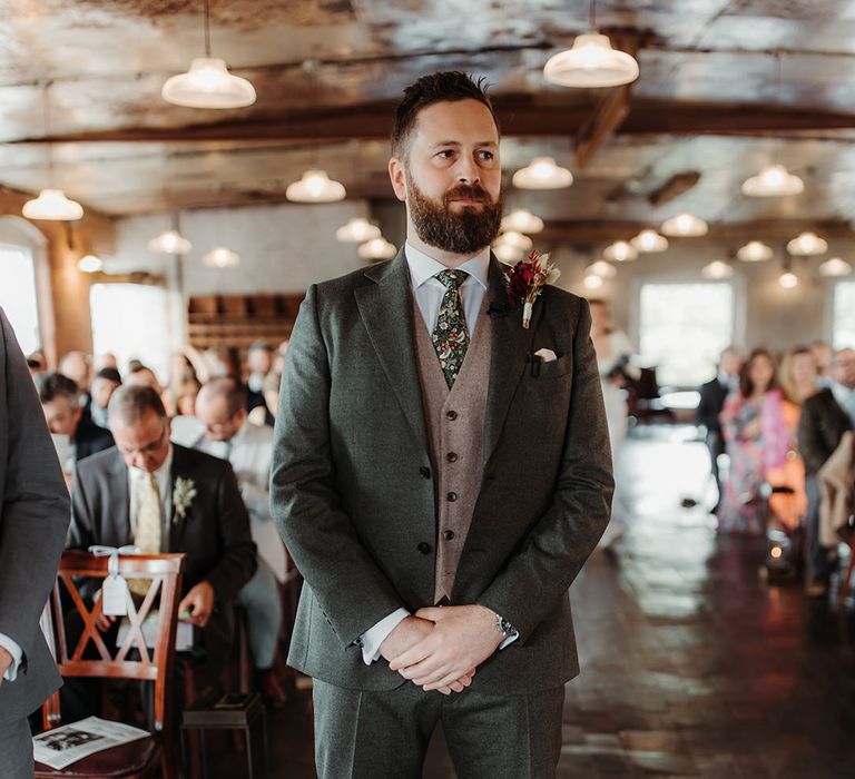 Groom in green wedding suit with grey waistcoat and floral patterned tie waiting for the bride at the end of the aisle 