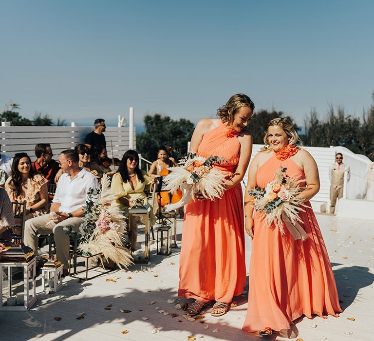 Bridesmaids in coral halterneck bridesmaid dresses walking down the aisle together 