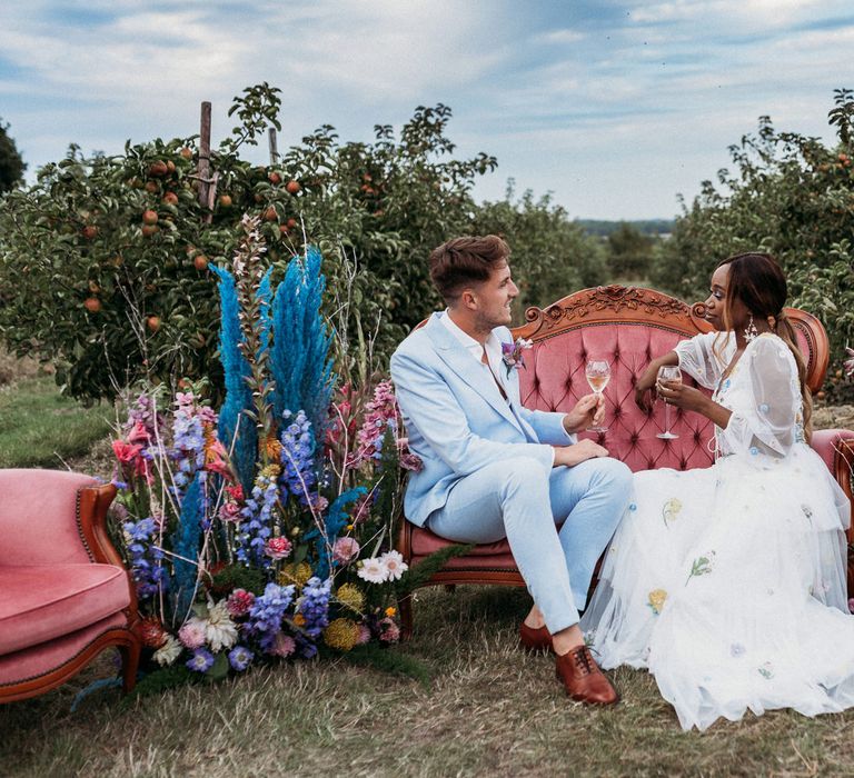 Bride and groom sitting on an ornate chair sipping champagne