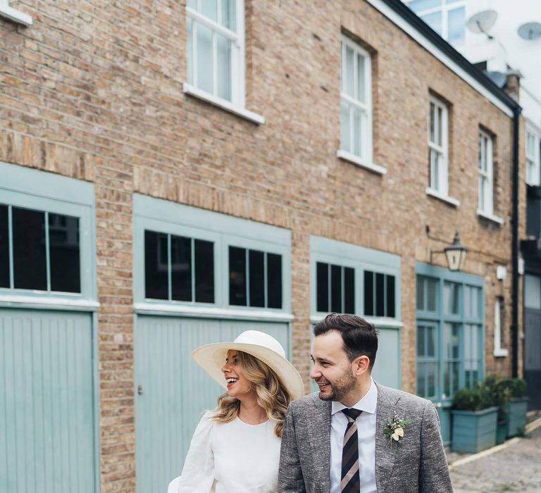 Bride and groom holding hands in Marylebone bride wears Net A Porter wedding dress and nude heels with wedding hat