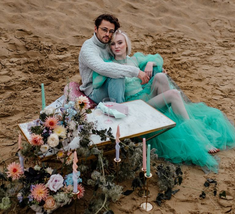 Bride in a pastel green wedding dress sitting on the sand by her picnic table decorated with colourful flowers and candles with her groom in a grey jumper 