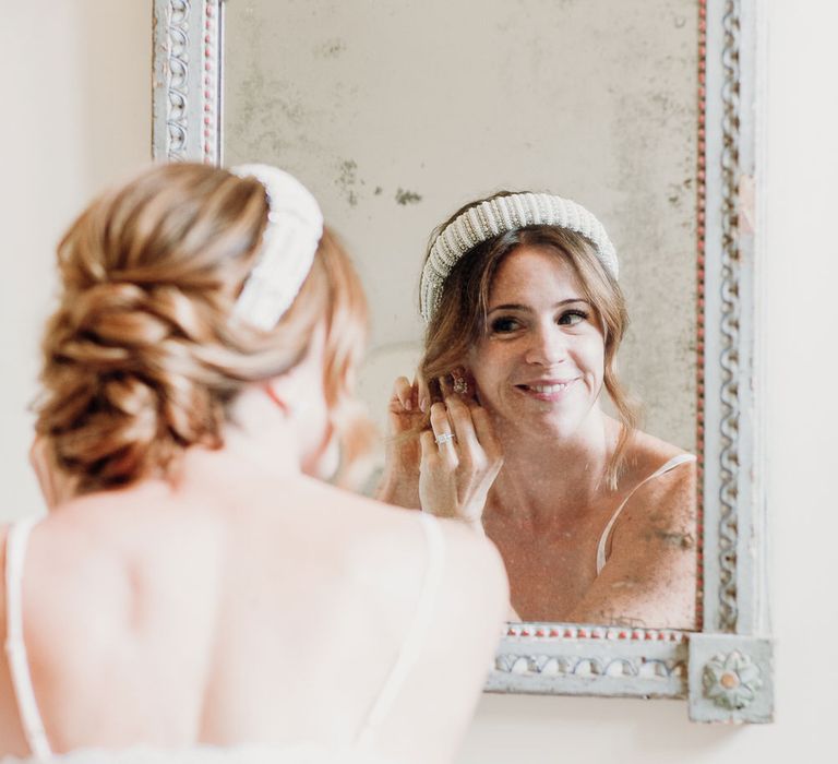 Bride with wedding up-do and beaded bridal headband smiles into mirror whilst putting on earrings 