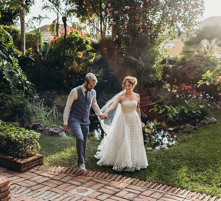 Groom in a light blue waistcoat holding hands with his bride in a strapless wedding dress at Gibraltar Botanic Gardens