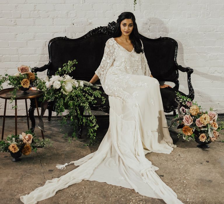 Bride in an embellished wedding dress sitting on a sofa surrounded by pink and green wedding floral arrangements 