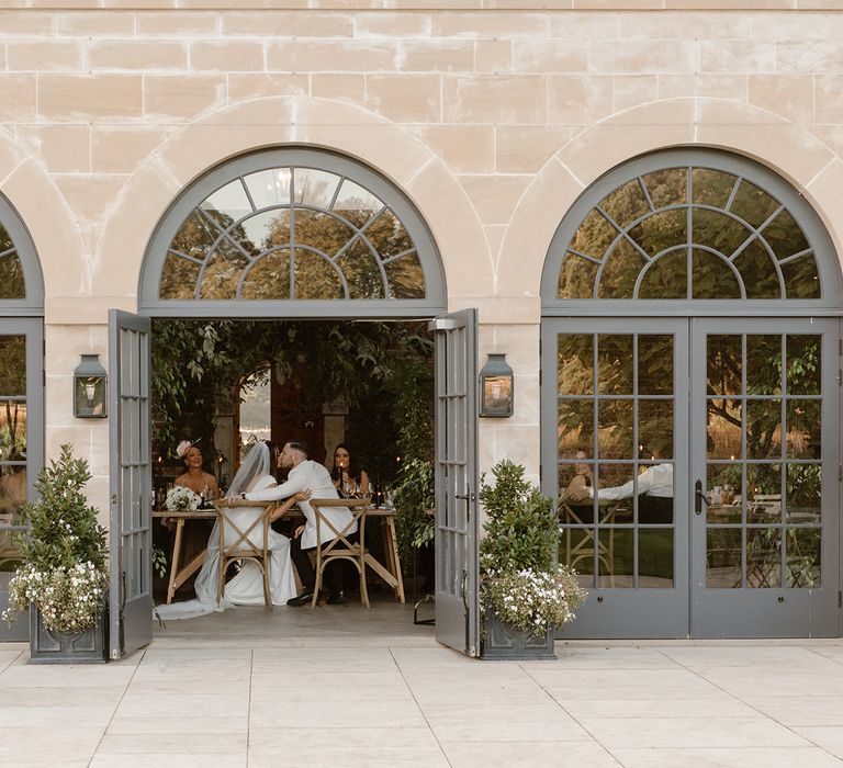 Bride and groom embrace sat at wedding table through open doors in the Fig House at Middleton Lodge