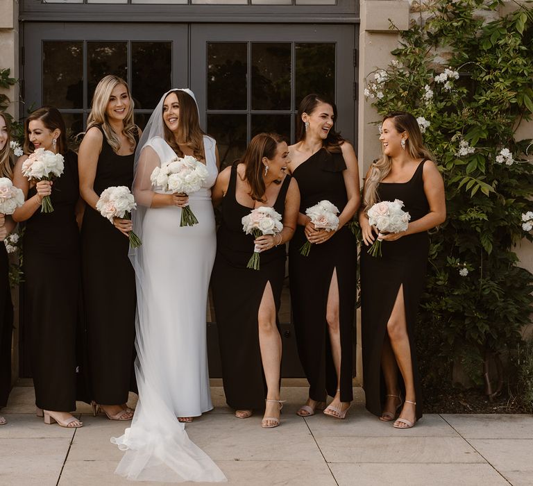 Bride in white Made With Love wedding dress poses outside with bridesmaids in long black bridesmaid dresses all holding white and blush pink rose bouquets