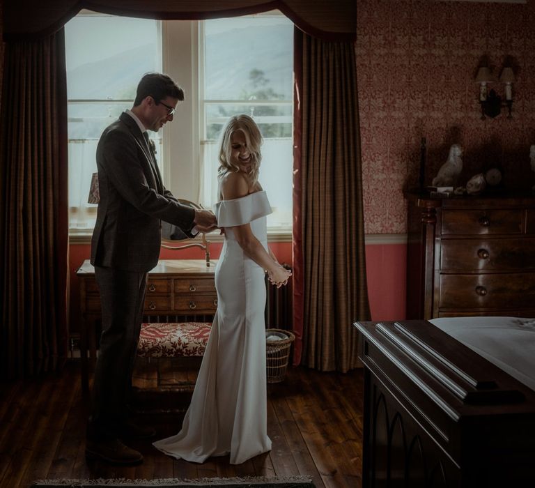 Groom helps bride with wedding dress on the morning of their wedding