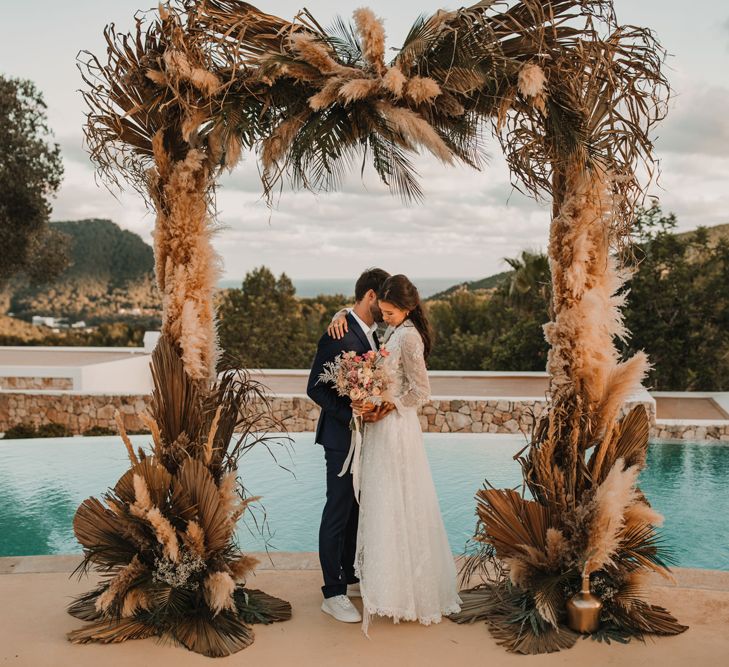 Bride and groom embracing under a dried grass, palm leaves and pampas floral arch 