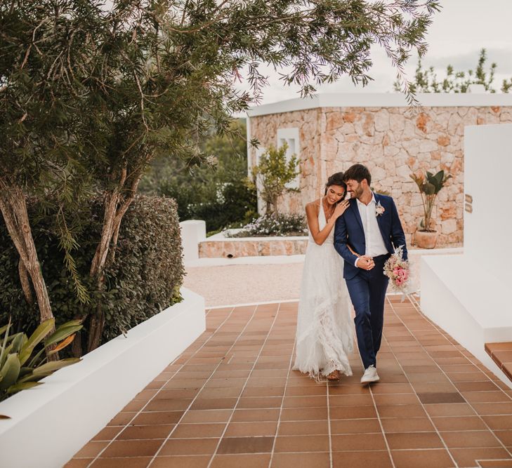 Boho bride in a halterneck Marylise Bridal gown with tassels holding her grooms hand in a navy suit 