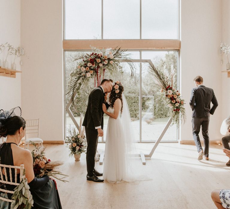 Boho ASOS wedding dress bride with tulle skirt and spaghetti straps kissing her husband at the wooden frame altar 