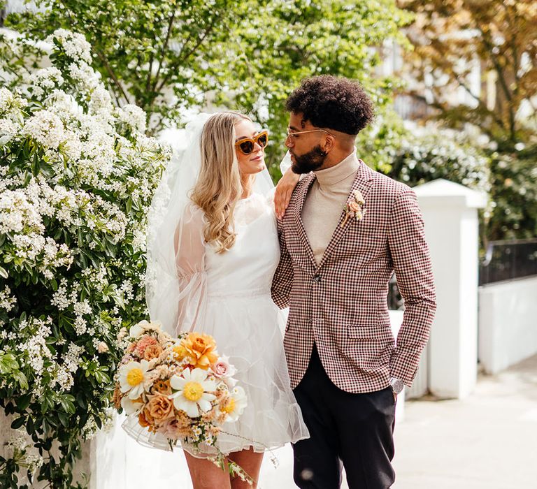 Stylish groom in a check blazer embracing his bride in a short ruffle wedding dress, sunglasses and platform boots holding a yellow wedding bouquet 