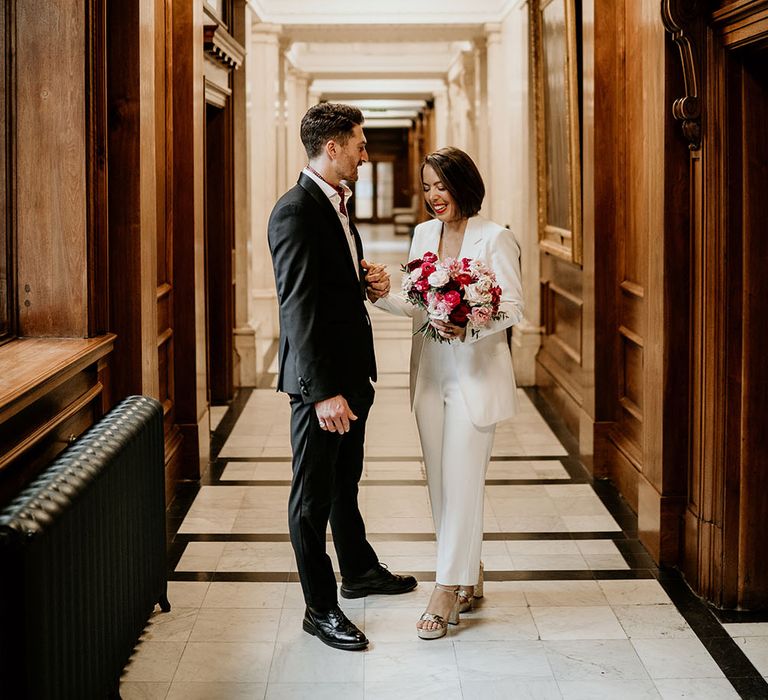 Bride & groom laugh together in the hallway as bride holds pink and red floral bouquet