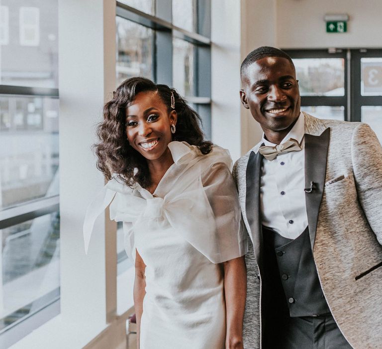 Smiling bride with short curled hair in white Bec + Bridge satin wedding dress with shoulder bow details holds hands with groom in grey suit jacket with black lapels and black trousers at Bridge Community Church wedding