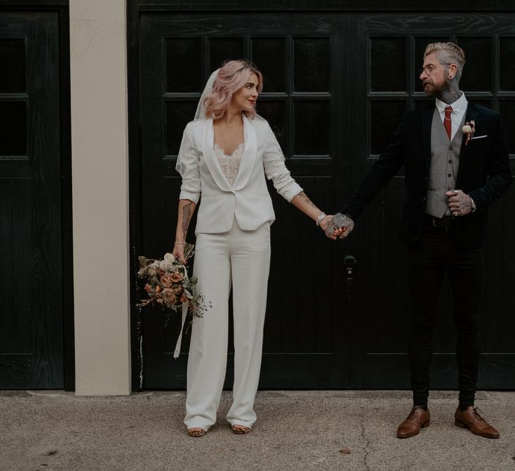 Bride in white suit and groom in dark suit stand side by side holding hands against black backdrop