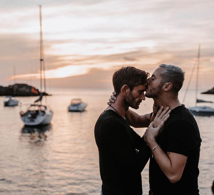 Groom kisses the forehead of his partner as the sea ripples behind them