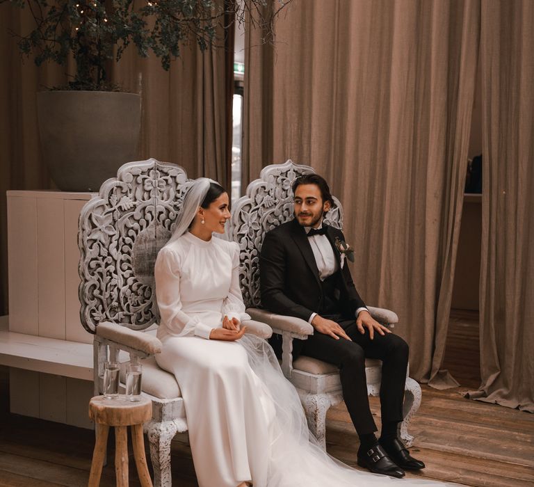 Bride & groom sit together on silver thrones whilst looking lovingly at one another