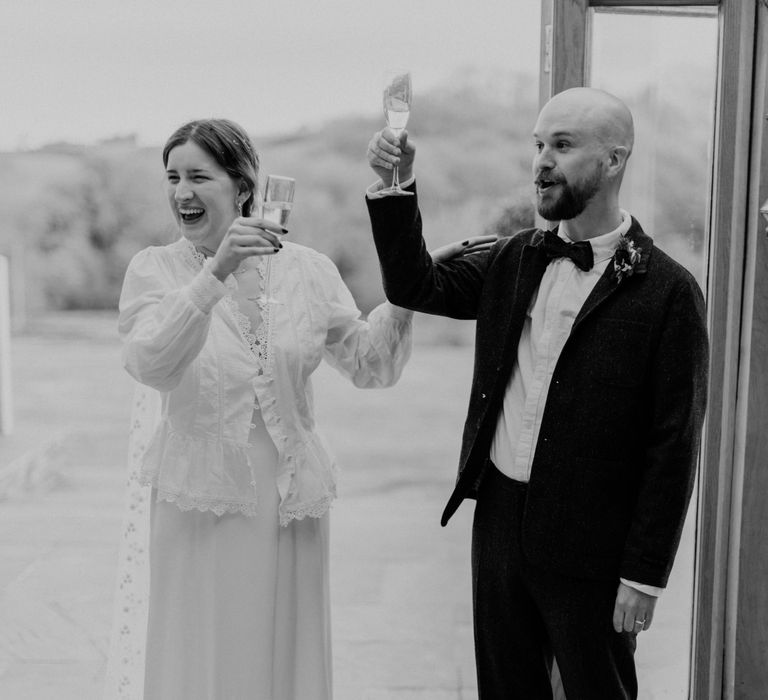 Smiling bride in white blouse, Charlie Brear wedding dress and chapel length daisy applique veil raises glass to guests with groom in grey tweed suit and bow tie at garden party wedding in Devon