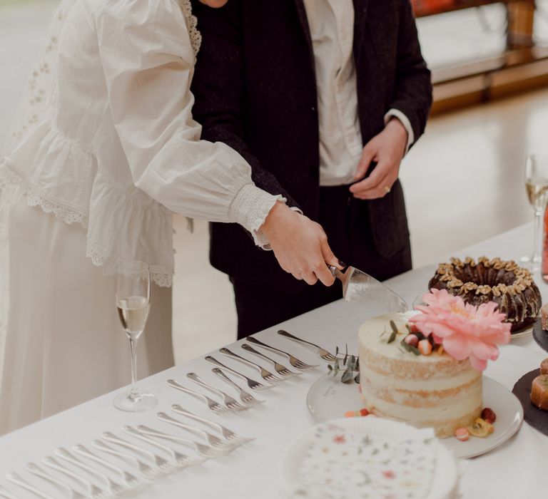 Smiling bride in white blouse, Charlie Brear wedding dress and applique veil cuts wedding cake with groom in grey tweed suit and bow tie at garden party wedding in Devon