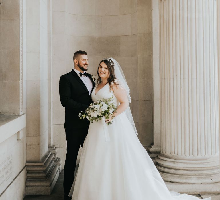 Bride & groom stand outside Old Marylebone Town Hall whilst bride holds white floral bouquet 