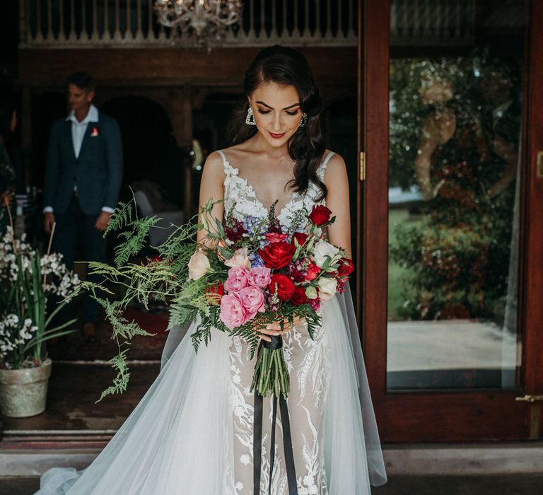 Bride in delicate lace embroidered wedding dress with layered tulle overskirt, holding bouquet of red and pink peonies, roses and ferns