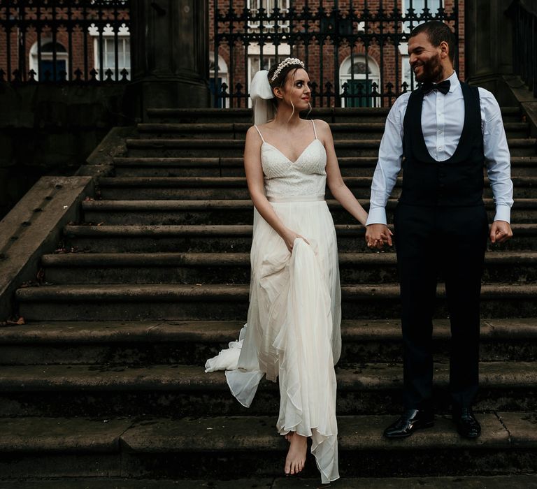 Groom in a horseshoe waistcoat and bow tie walking down the steps holding hands with his bride in a thin strap wedding dress and bow veil 