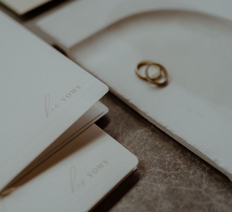 A booklet reading 'her vows'. Blurred in the corner of the shot are some wedding rings.