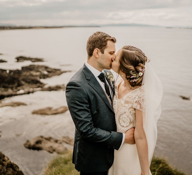 Bride in lace top wedding dress with satin skirt and veil kisses groom in navy suit on clifftop after Dunluce Castle wedding