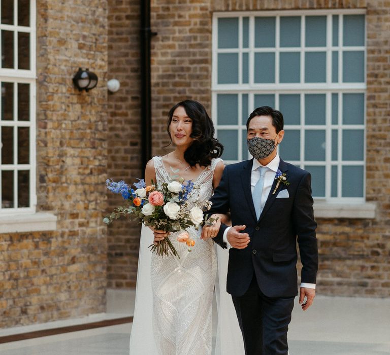 Bride walking down the aisle with her father, she is wearing an Eliza Jane Howell gown and carry a blue, peach and white bouquet