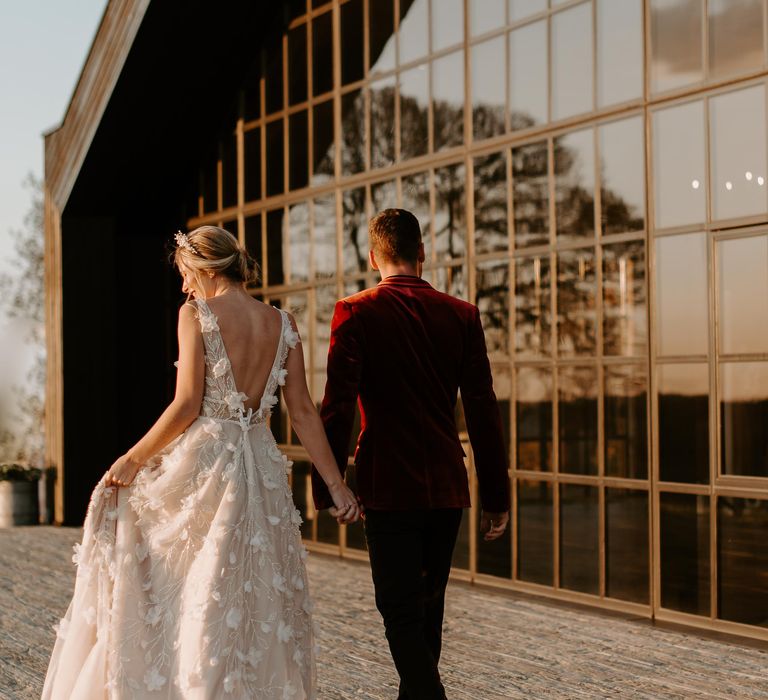 Bride in an appliqué wedding dress and groom in a red velvet tuxedo jacket walking in front of the windows at Botley Hill Barn 